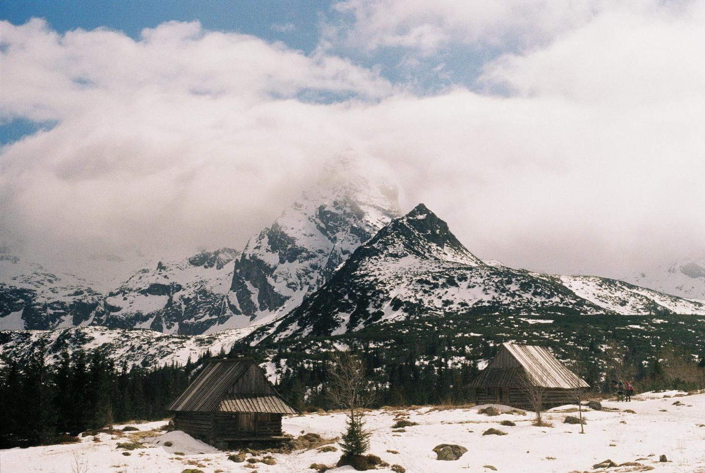 Two wooden cabins sit in a snowy landscape with mountains and clouds in the background.