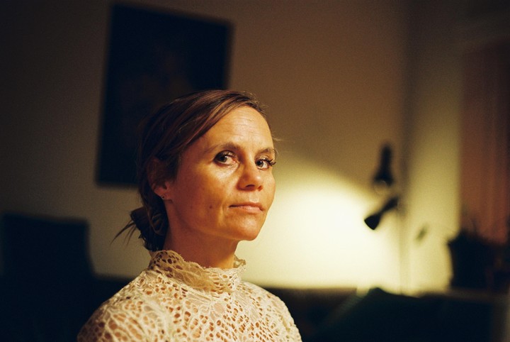 A woman with her hair tied back wears a high-necked lace top and looks directly at the viewer.