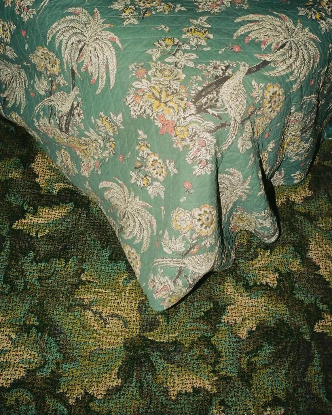 A green patterned quilt with palm trees and birds lies on a green patterned carpet.