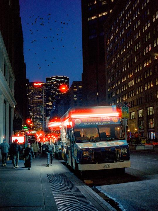 A food truck with bright red lights is parked on a city street at night, with people walking on the sidewalk and tall buildings in the background.
