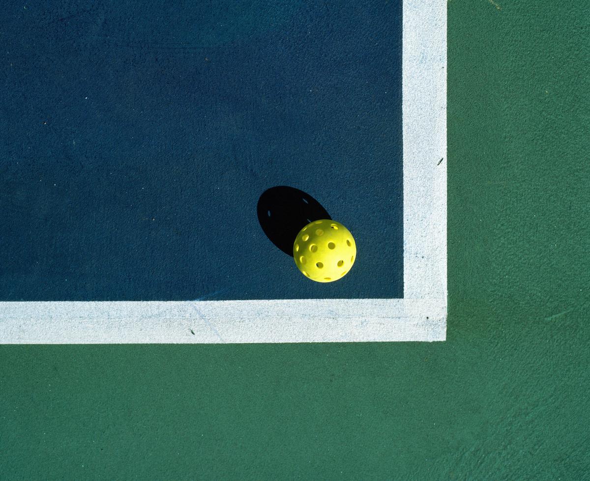 A yellow pickleball with a shadow sits on the corner of a blue and green court with white lines.