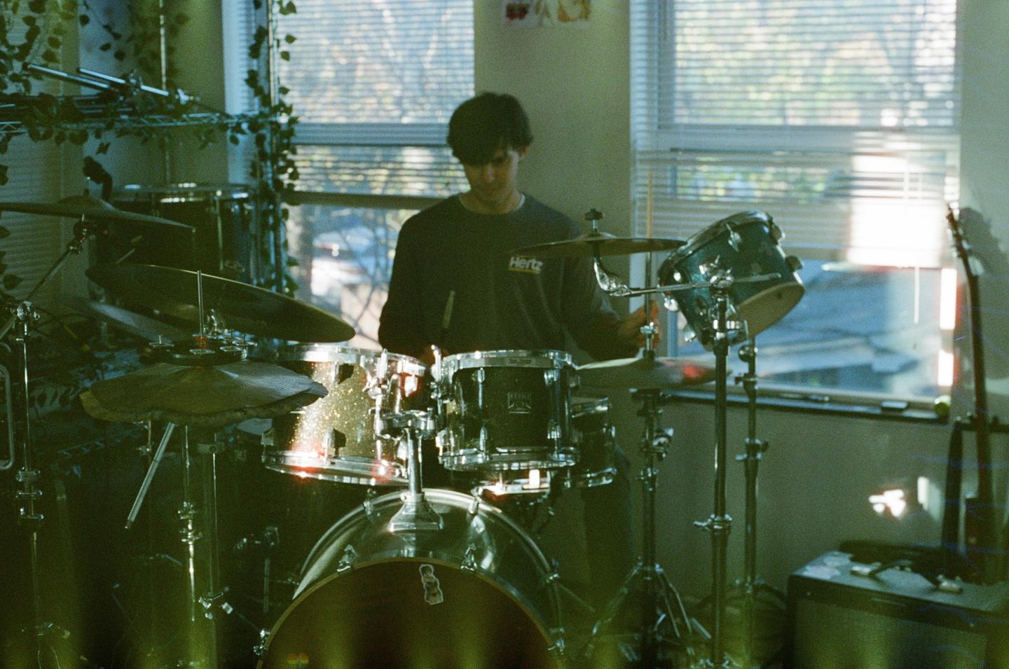 A man sits behind a drum kit in a room with a window.