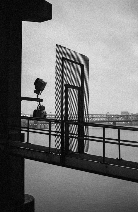 A black and white photograph shows a metal walkway with a railing and a mesh gate, with a body of water and a bridge in the background.
