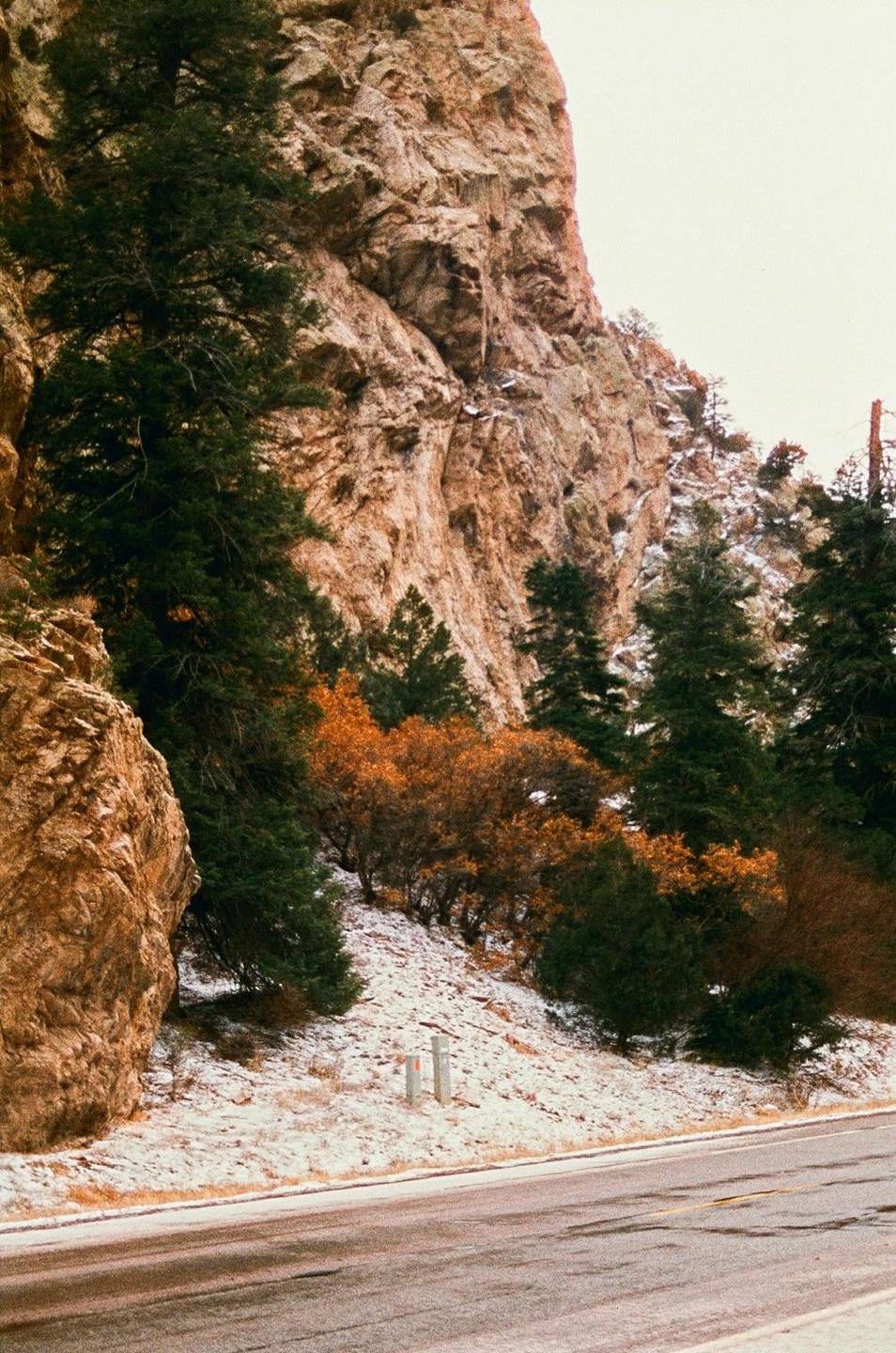A road runs alongside a snow-dusted hill with evergreen and autumn trees, next to a large rock formation.