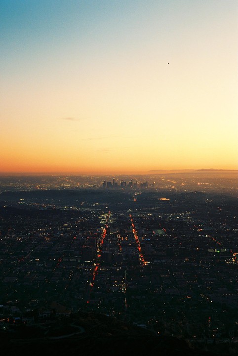 A city with illuminated streets and buildings stretches into the distance under a gradient sky at dusk.