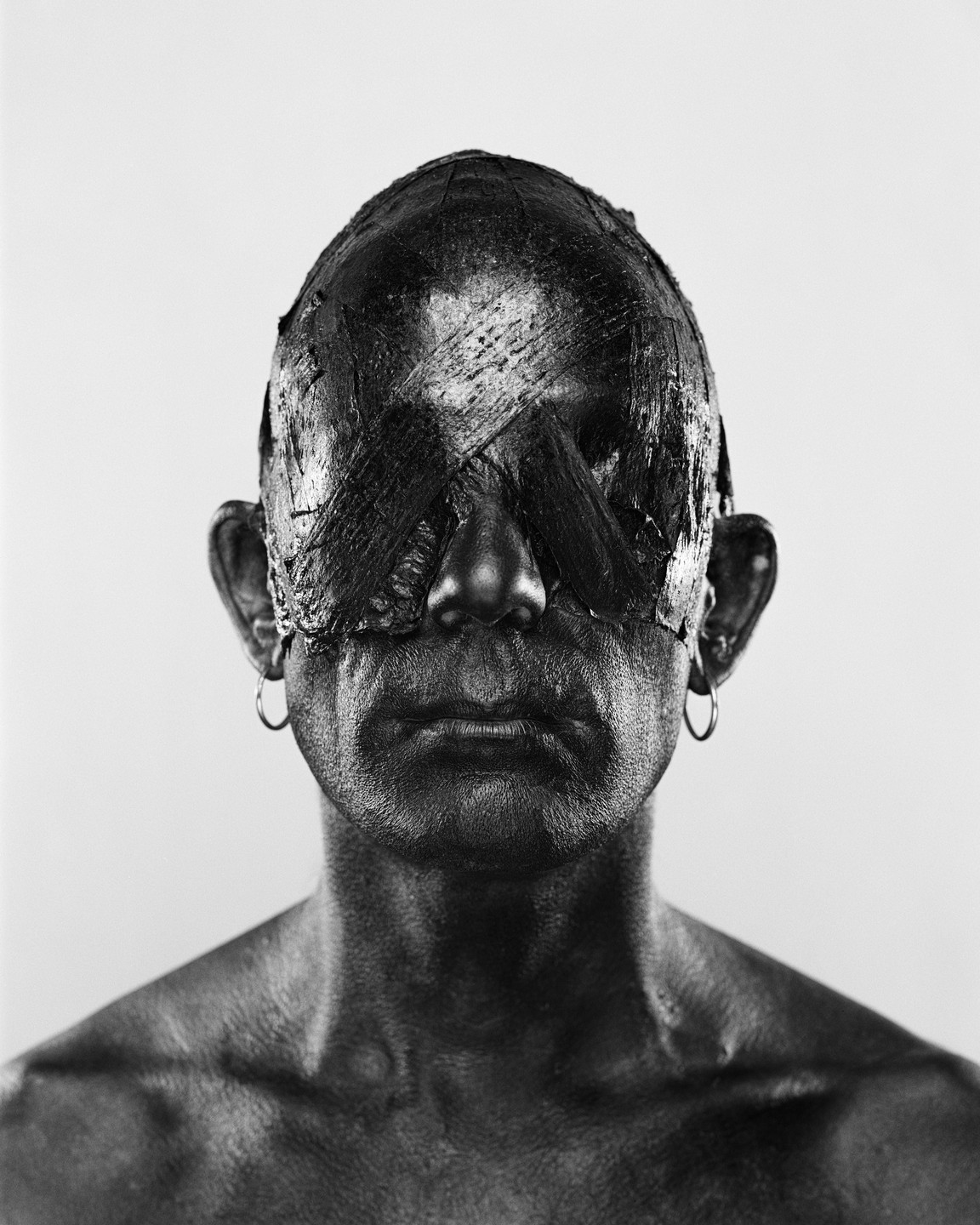 A man with dark paint covering his head and eyes, wearing hoop earrings, looks directly forward.