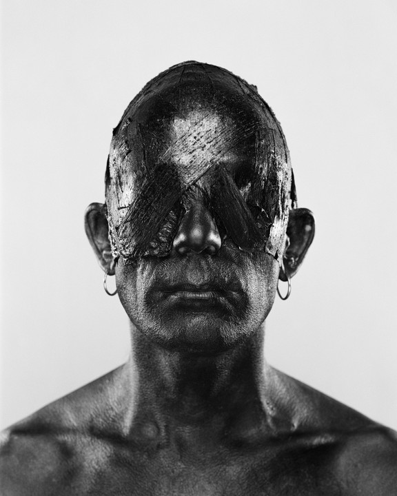 A man with dark paint covering his head and eyes, wearing hoop earrings, looks directly forward.