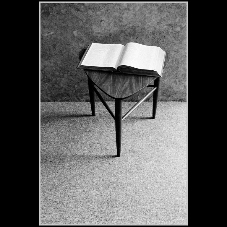 An open book rests on a small wooden table in a room with a textured wall and carpeted floor.