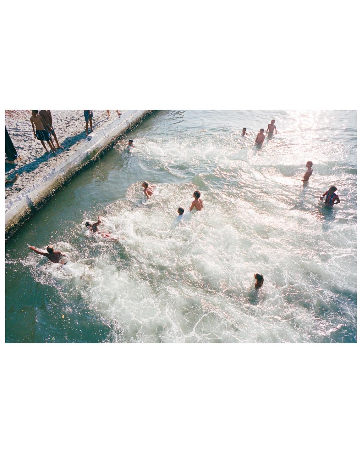 A group of children and adults are swimming and playing in the foamy water next to a sandy bank.