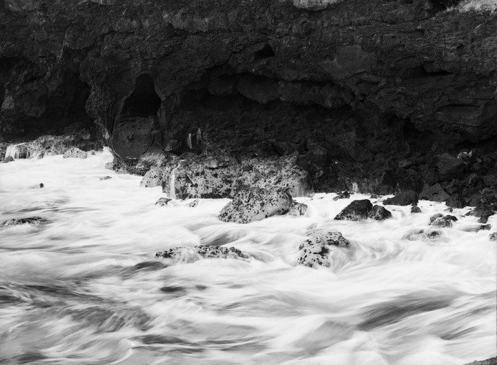 Water rushes around rocks at the base of a dark cliff with a cave opening.