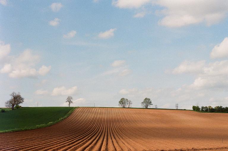 A vast landscape shows a brown plowed field in the foreground and a green hill with trees in the background under a blue sky with clouds.