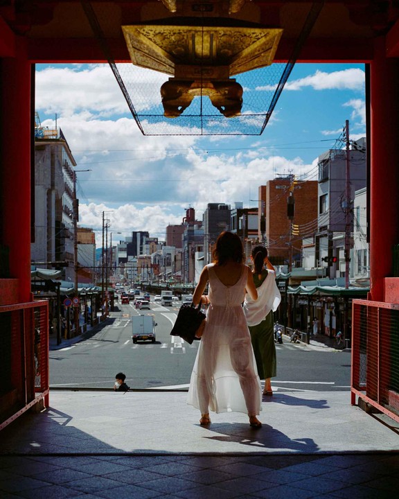 Two women stand at the entrance of a building looking out at a city street with cars and buildings under a blue sky with clouds.