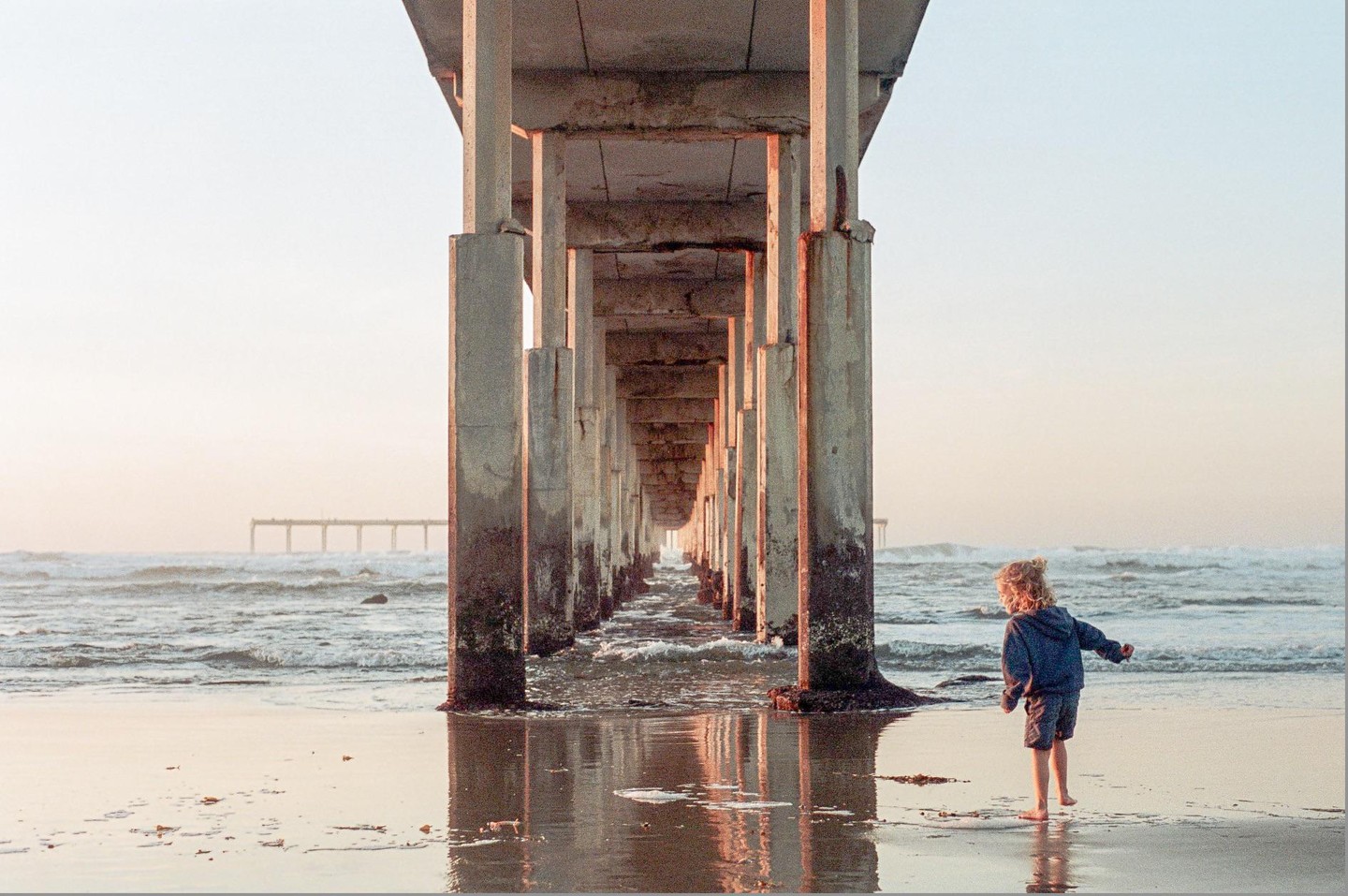 A child stands barefoot on a wet beach under a pier with waves in the background.