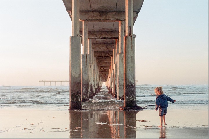 A child stands barefoot on a wet beach under a pier with waves in the background.