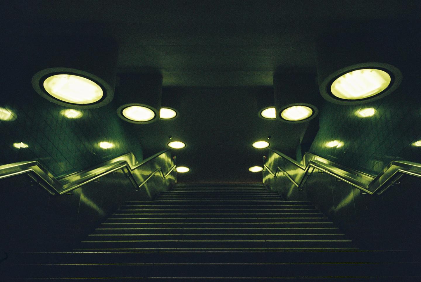 A long, dark staircase with bright, circular lights on the ceiling and walls leads upwards.