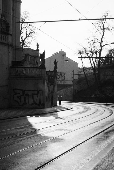A man walks along a curved street with tram tracks and a building with statues and graffiti.