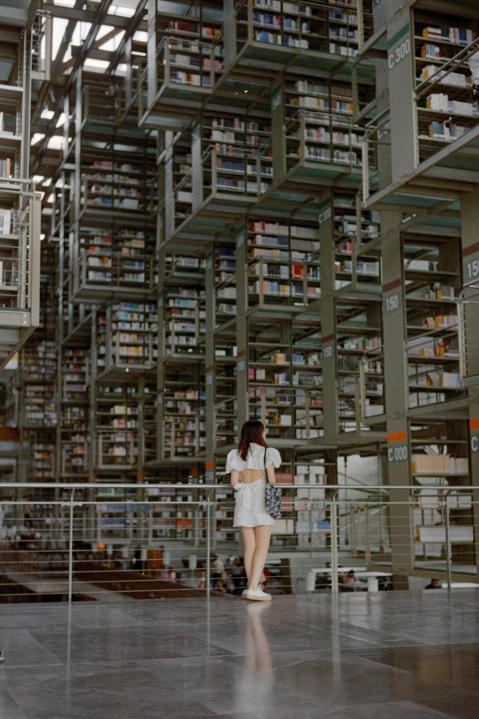 A woman stands on a balcony overlooking a vast library with towering bookshelves.