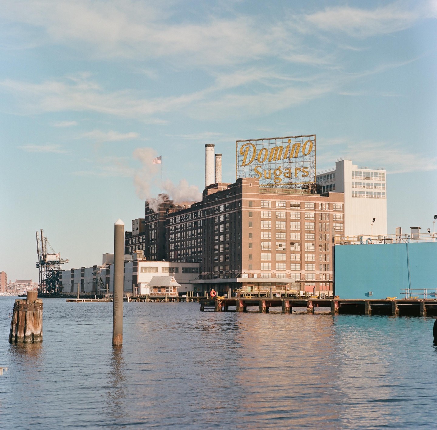 A large building with a "Domino Sugars" sign on its roof stands next to a body of water with a crane and other structures in the background.