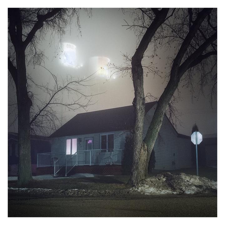A house with a glowing window is seen through bare trees on a foggy night with illuminated water towers in the background.