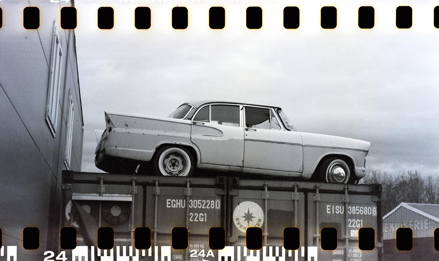 A vintage car is parked on top of a shipping container next to a building under a cloudy sky.