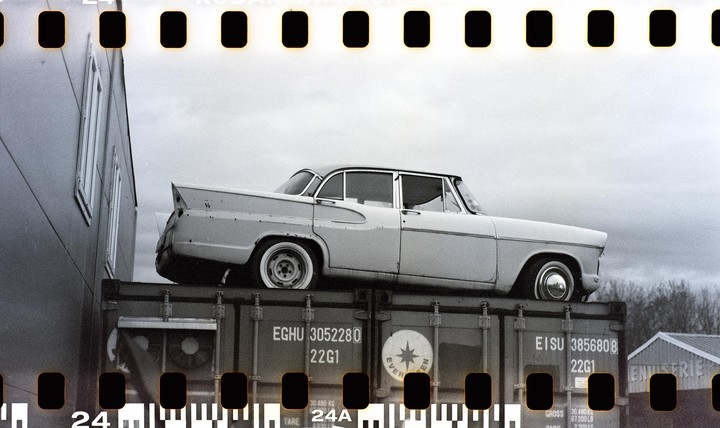 A vintage car is parked on top of a shipping container next to a building under a cloudy sky.