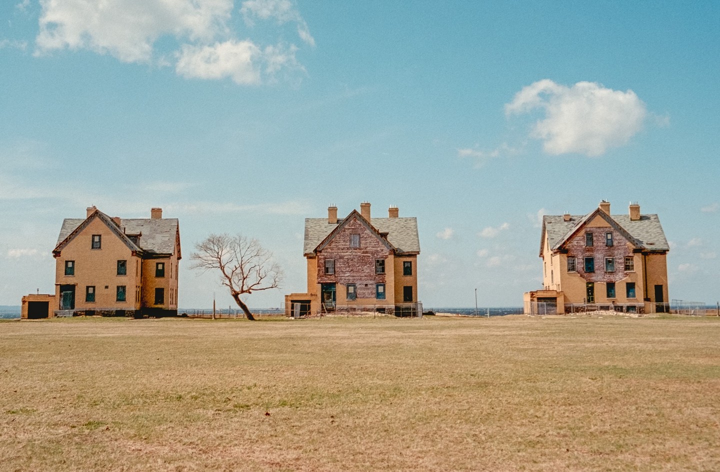 Three large, old houses stand in a field with a bare tree next to one of them, under a blue sky with scattered clouds.