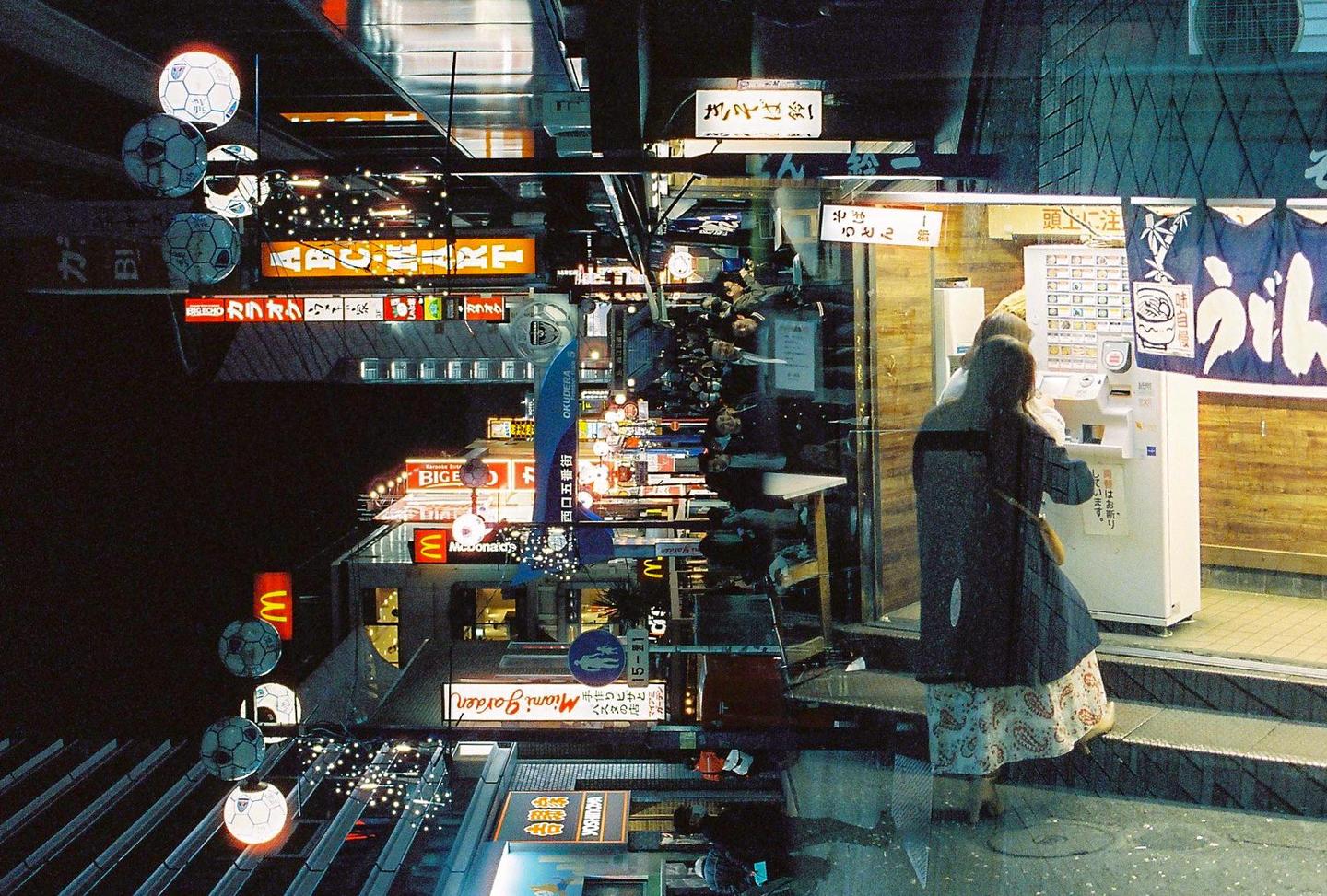 A woman stands at a vending machine on a city street at night, with reflections of neon signs and buildings visible in the foreground.