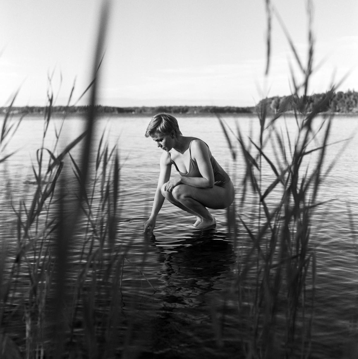 A woman in a swimsuit crouches in a lake and touches the water with her hand.