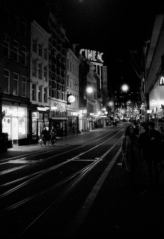 A street at night is lined with buildings and illuminated signs, with people walking and cycling.