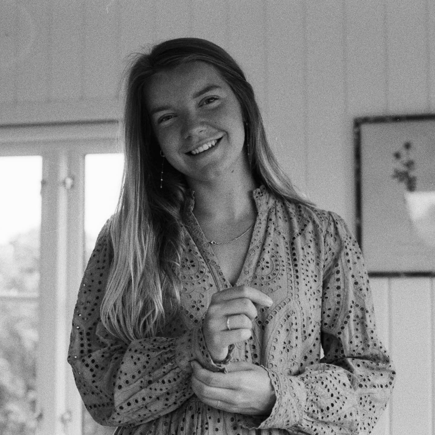 A smiling woman with long hair wears a patterned dress and jewelry while standing indoors.