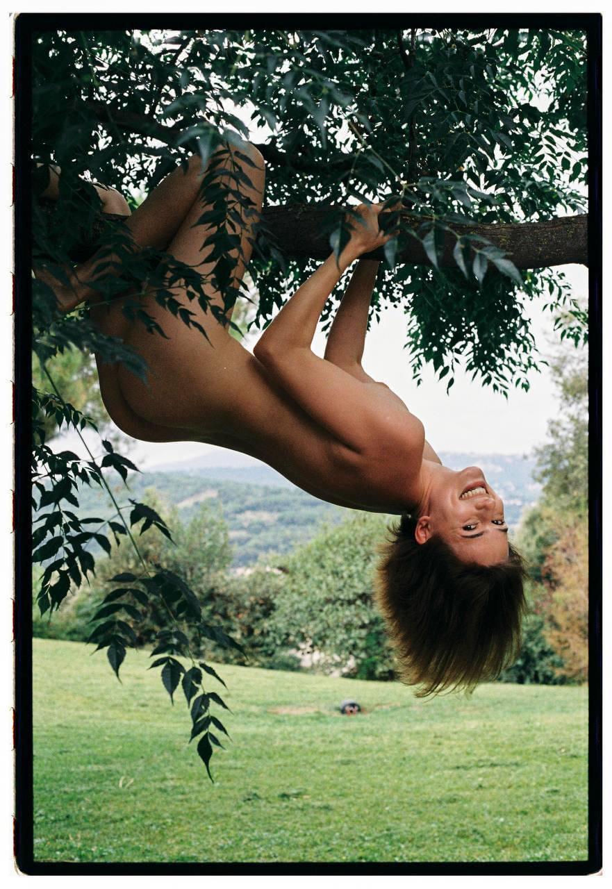A woman hangs upside down from a tree branch, smiling at the camera.