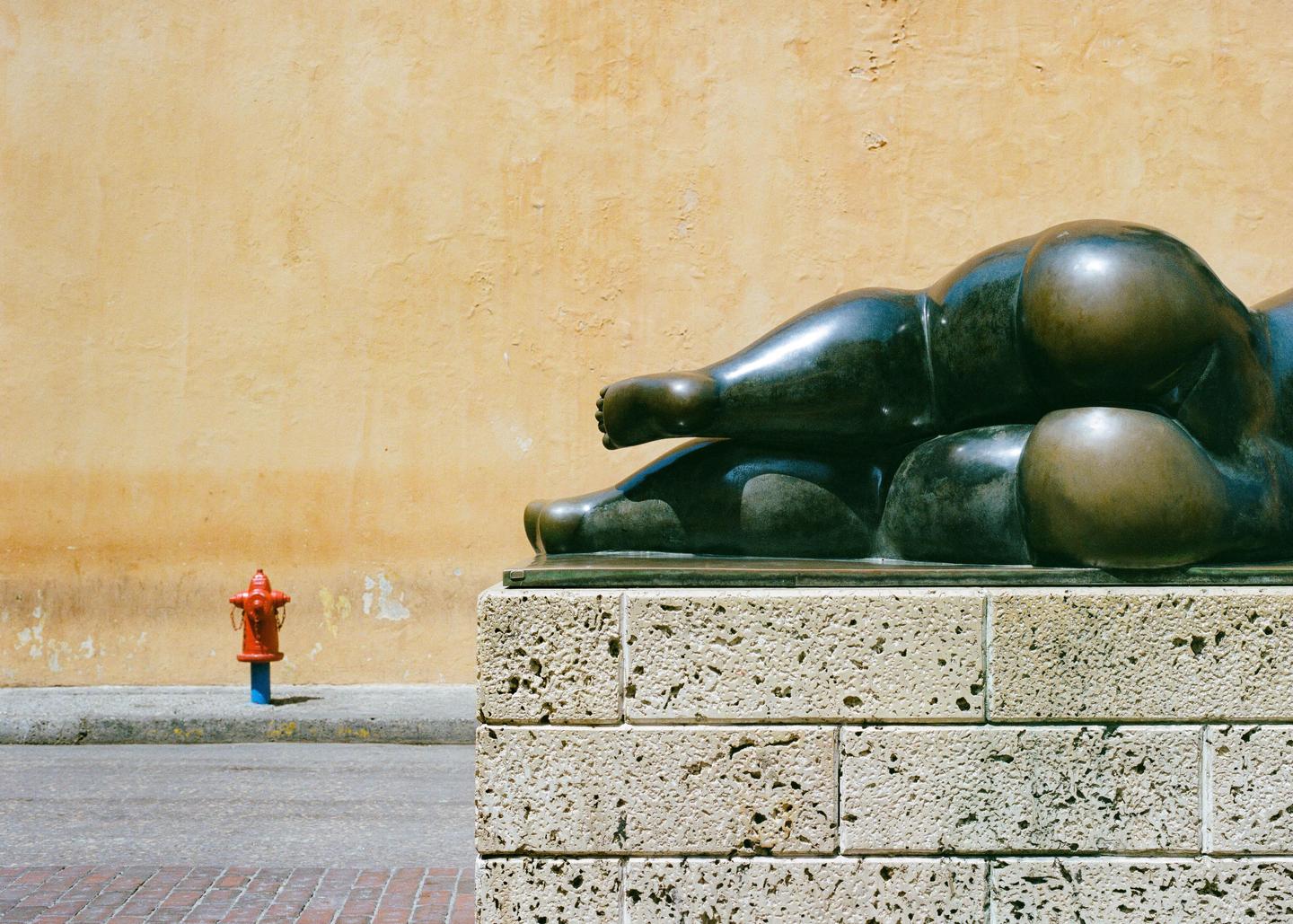 A bronze sculpture of a reclining figure rests on a stone base next to a street with a red fire hydrant.