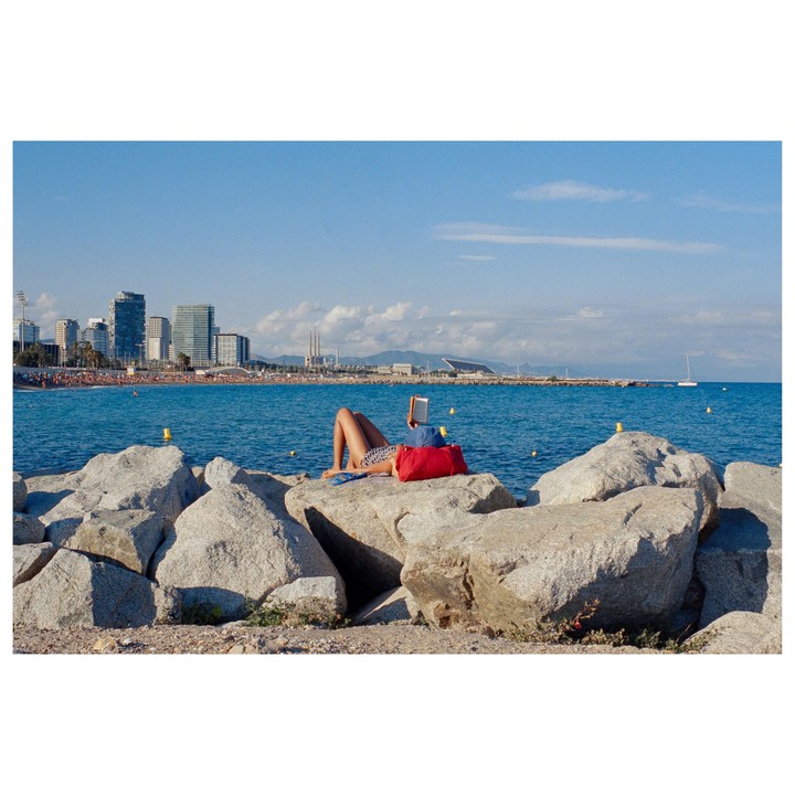 A woman in a swimsuit lies on a rock reading a book by the sea, with a city skyline in the background.