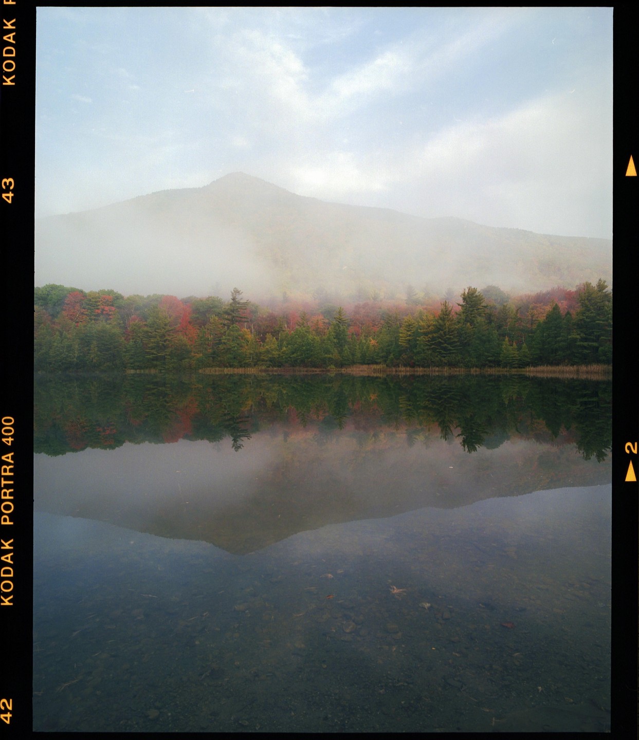 A foggy mountain rises above a lake reflecting autumn trees under a cloudy sky.