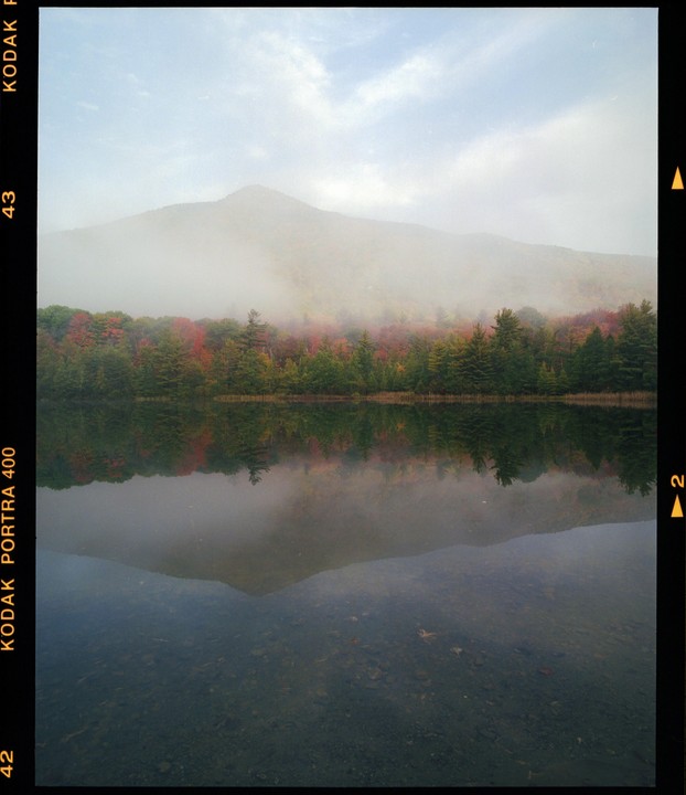 A foggy mountain rises above a lake reflecting autumn trees under a cloudy sky.