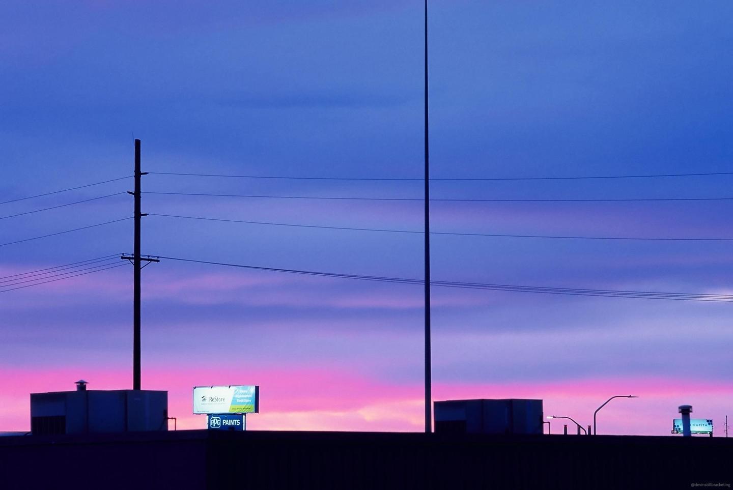 A vibrant pink and blue sunset sky is visible above a dark silhouette of buildings, power lines, and billboards.