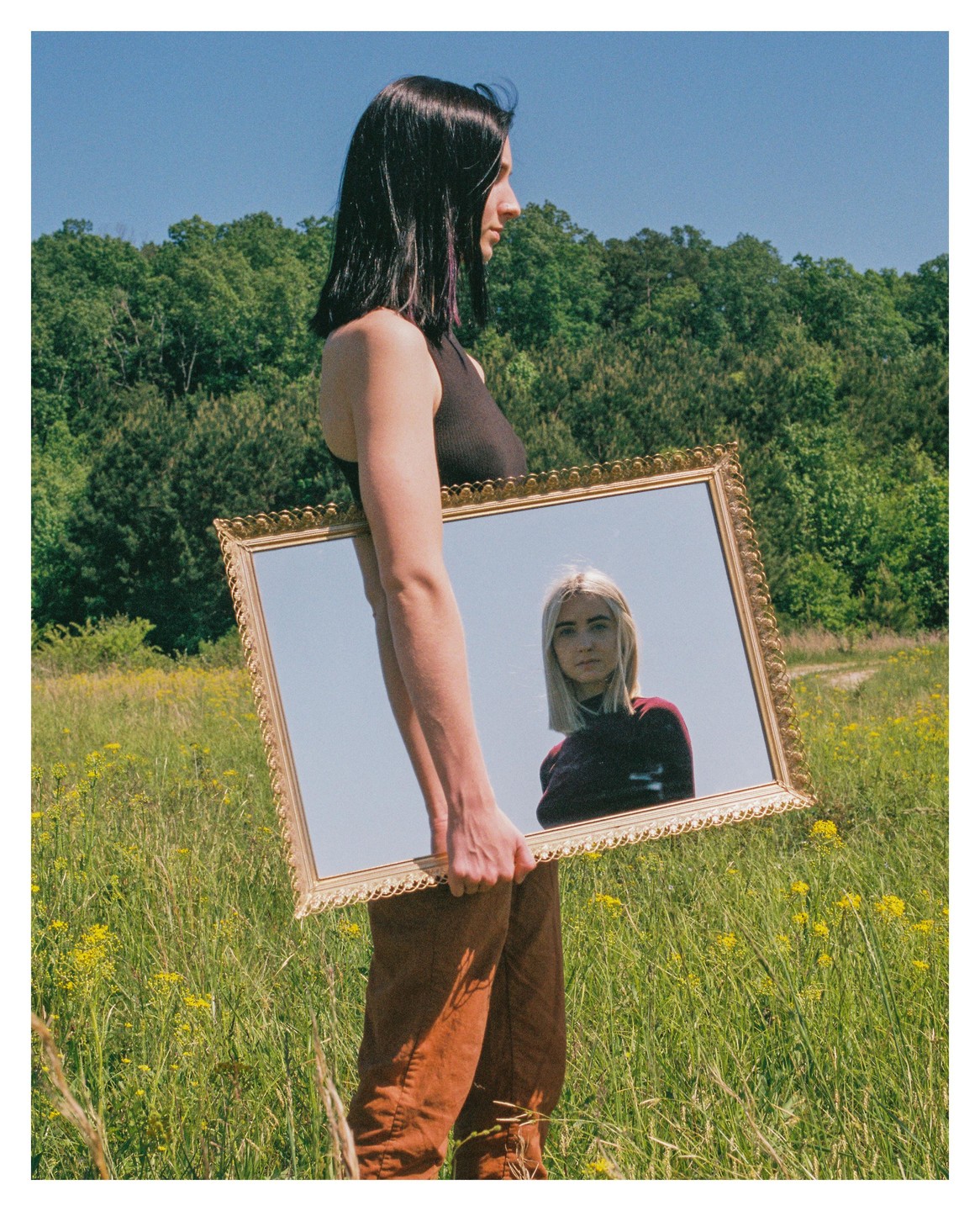 A woman with dark hair holds a large ornate mirror reflecting a blonde woman in a field of wildflowers.
