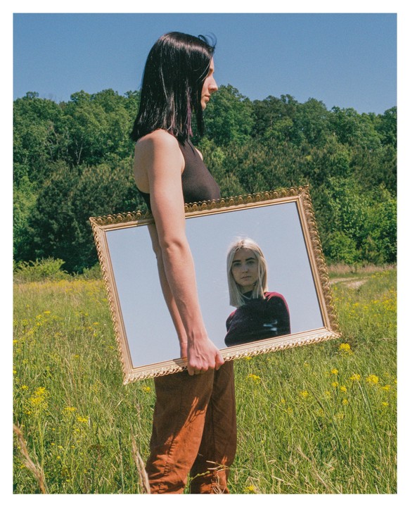 A woman with dark hair holds a large ornate mirror reflecting a blonde woman in a field of wildflowers.