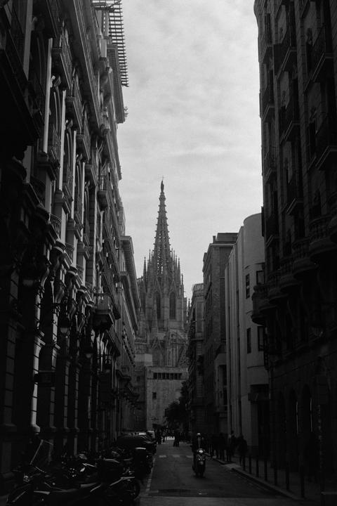 A narrow street with buildings on both sides leads to a tall, ornate church spire in the distance.