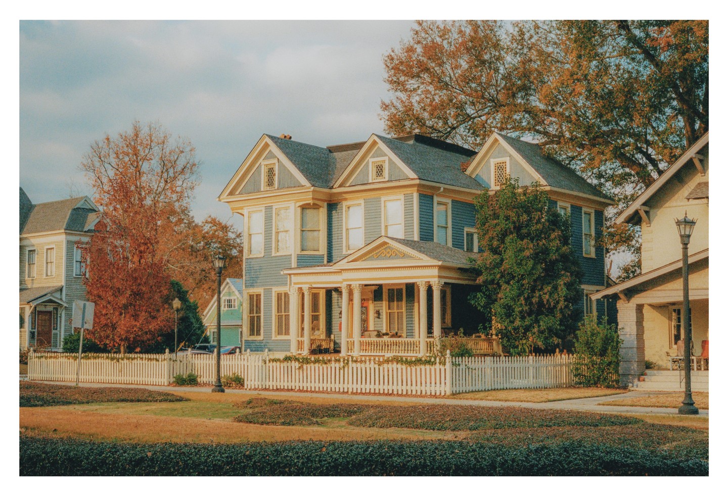 A large blue house with a white picket fence stands on a corner with autumn trees in the background.
