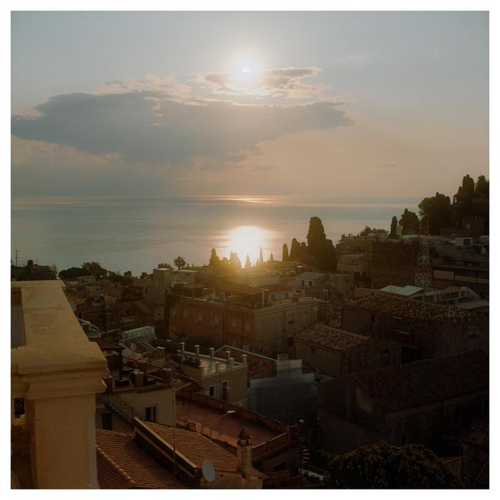 The sun sets over a town on a hill overlooking the sea, with a bright reflection on the water.