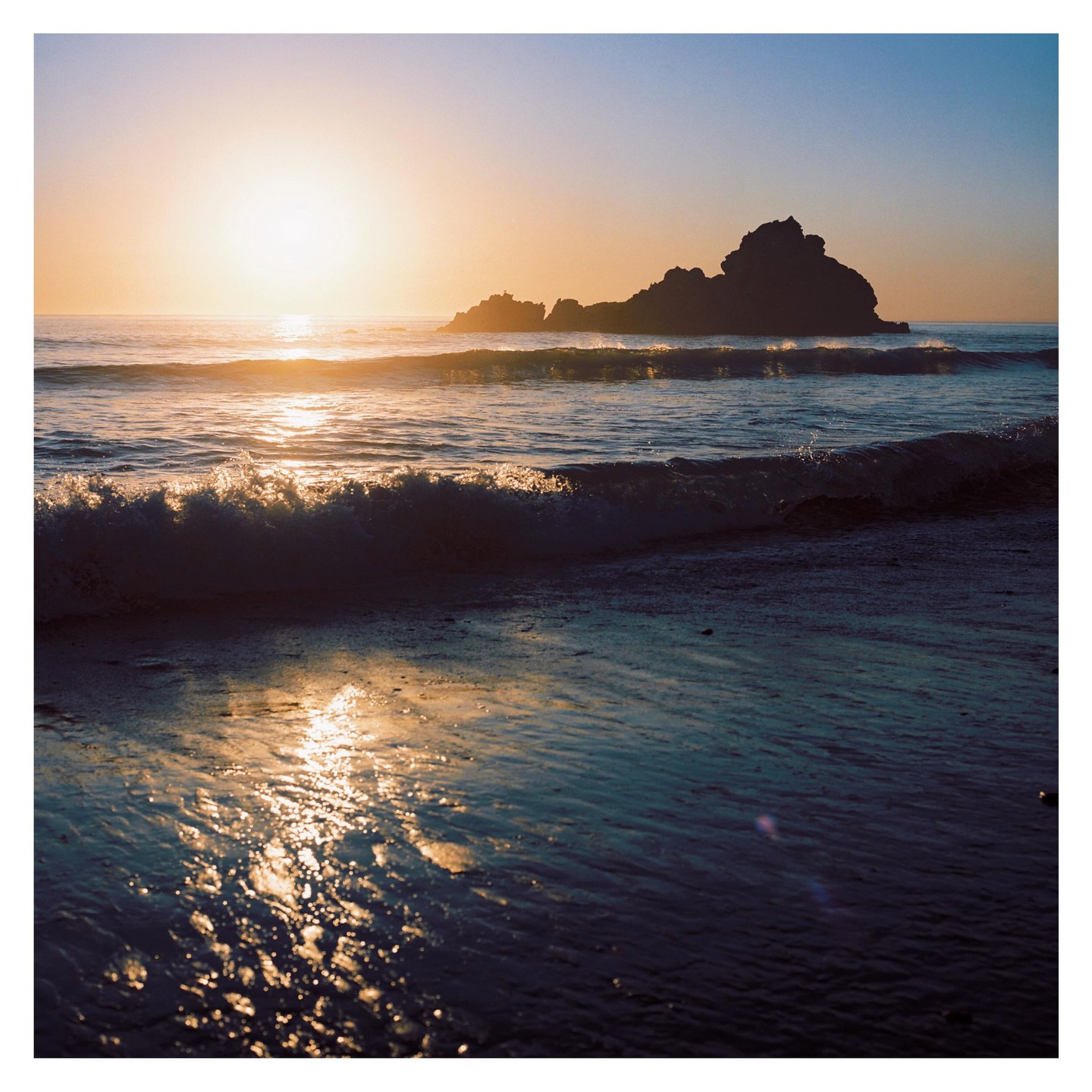 The sun sets over the ocean with waves crashing on the shore and a rocky island in the distance.