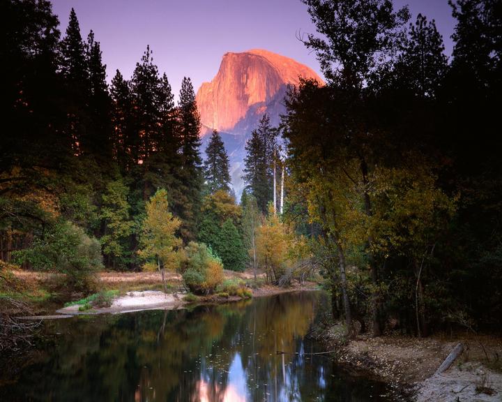 A river flows through a forest with a large mountain in the background at sunset.