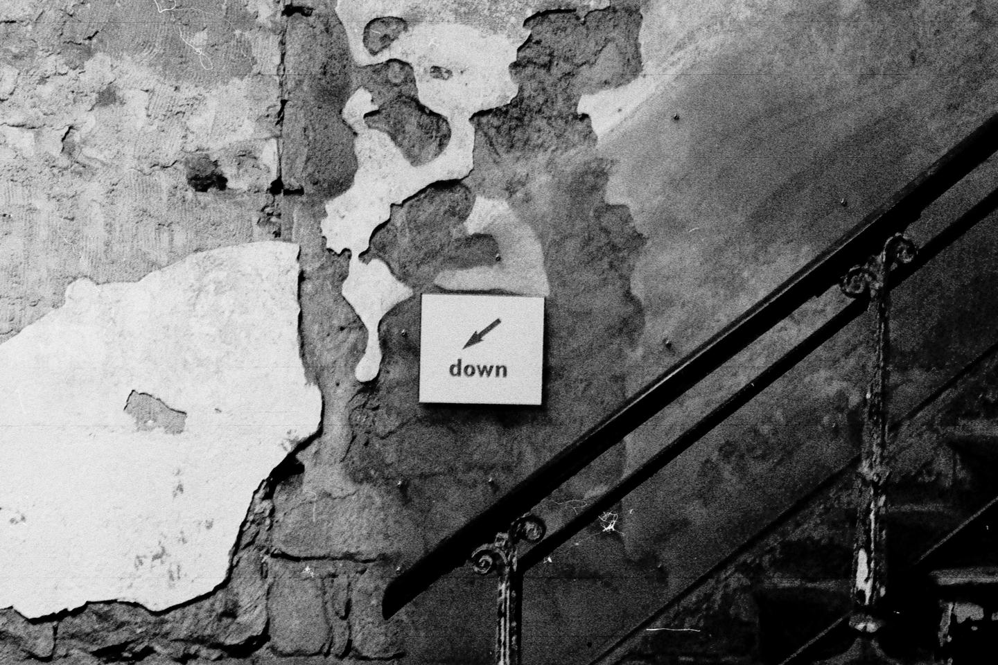 A sign with an arrow pointing down and the word "down" is affixed to a peeling wall next to a metal railing.