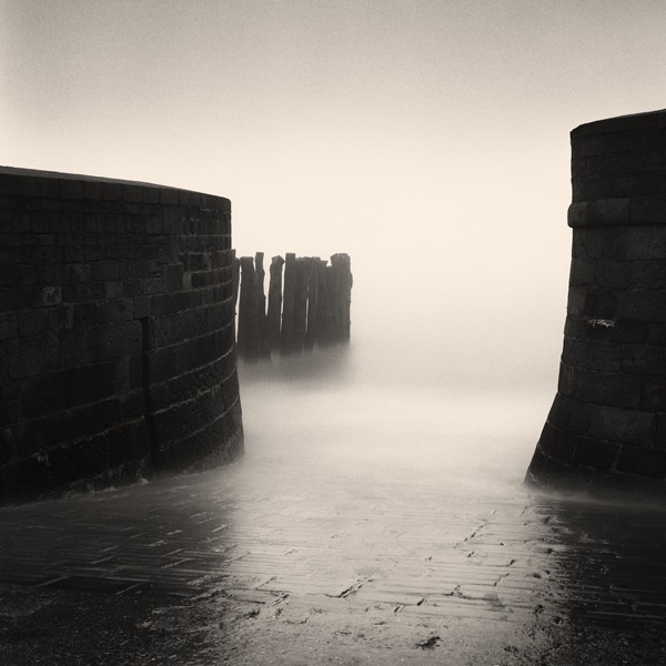 Two stone walls frame a misty body of water with wooden pilings in the distance.