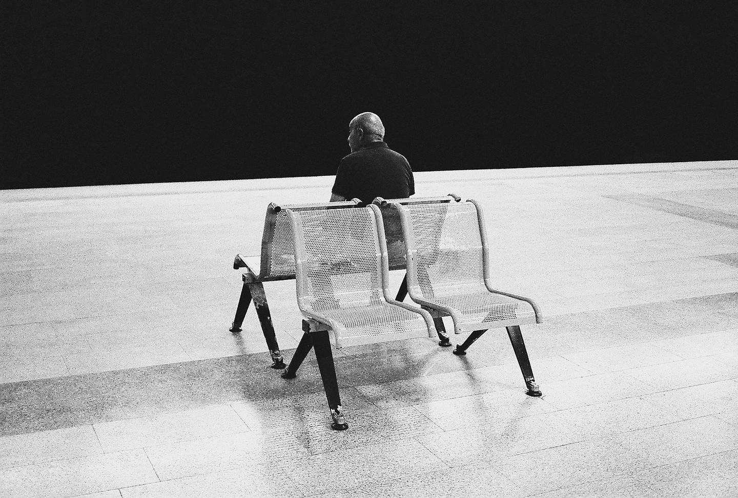 A man sits on a metal bench with his back to the viewer, facing a dark wall.