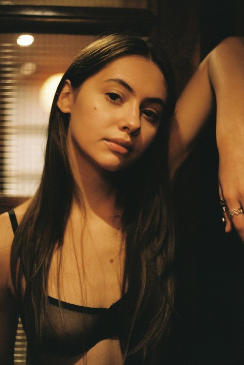 A young woman with long hair wears a sheer bra and looks directly at the viewer.