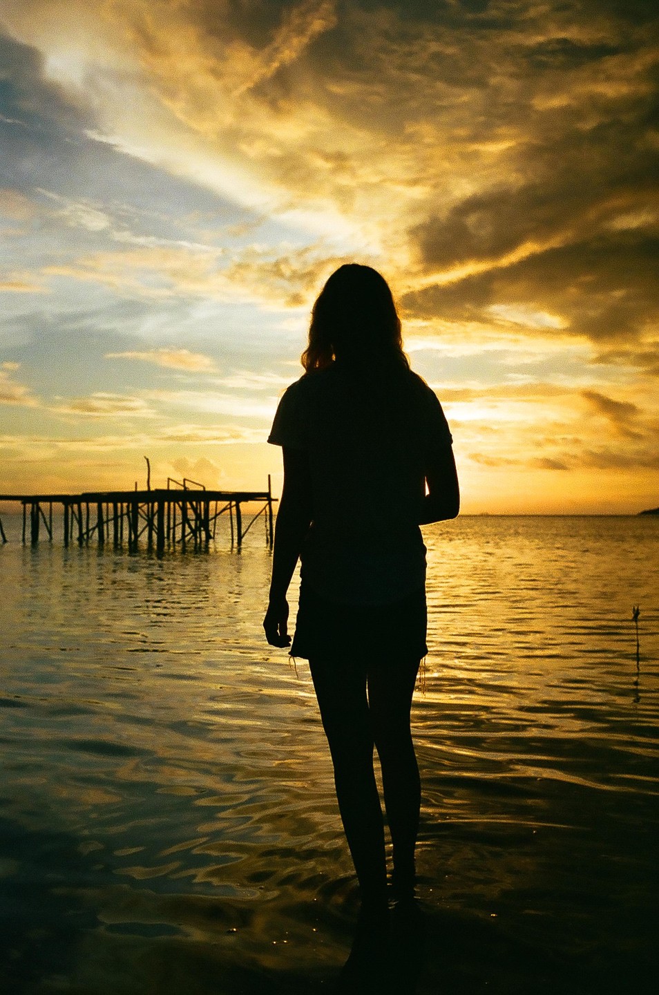 A woman stands in the water with a pier in the background at sunset.