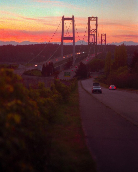 A suspension bridge with cars driving on it is visible at dusk, with a vibrant orange and blue sky in the background.