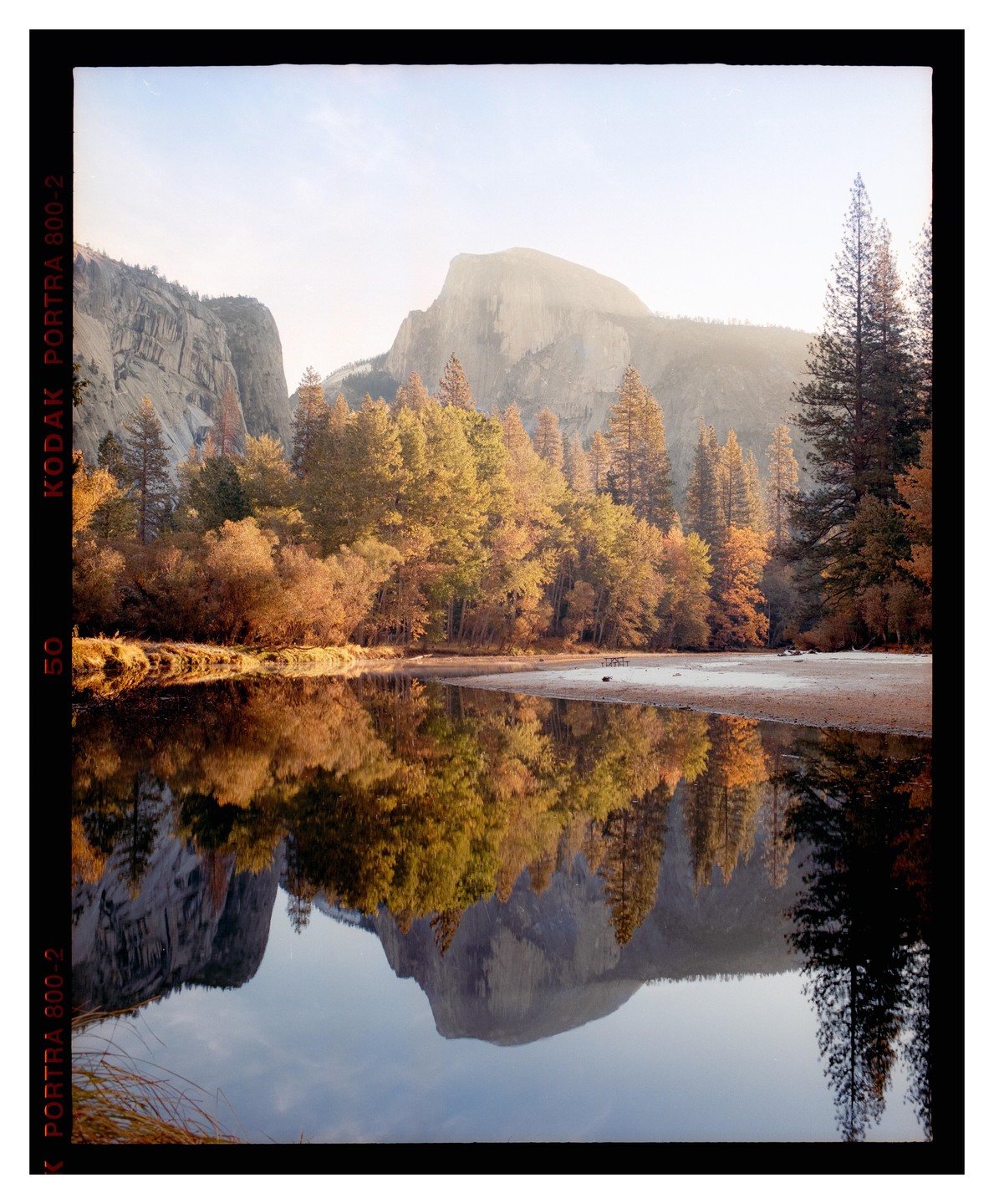 Half Dome mountain is reflected in a still river surrounded by autumn trees under a bright sky.